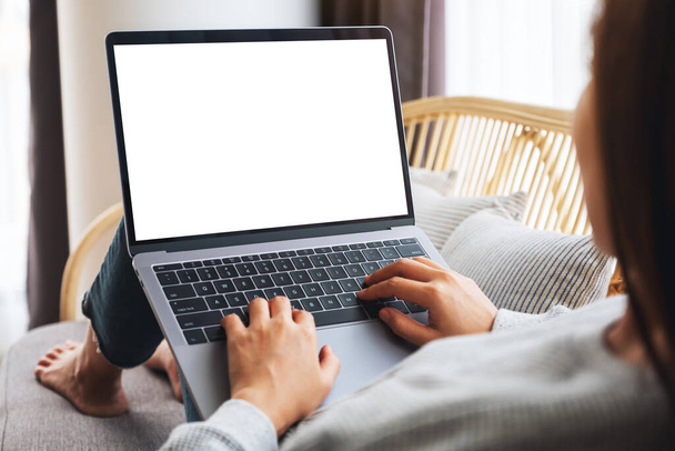 Mockup beeld van een vrouw die werkt en typt op laptop computer met leeg scherm tijdens het zitten op een bank thuis - Foto, afbeelding