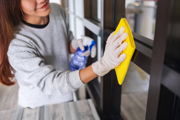 Μια νεαρή γυναίκα που φοράει προστατευτικό γάντι καθαρίζει το παράθυρο για την έννοια της οικιακής εργασίας - Φωτογραφία, εικόνα