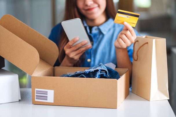 Close-up beeld van een mooie jonge vrouw met behulp van mobiele telefoon en creditcard voor online winkelen met boodschappentas en postpakket doos met kleding op de tafel - Foto, afbeelding