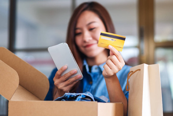 Nahaufnahme einer schönen jungen Frau, die Handy und Kreditkarte für den Online-Einkauf mit Einkaufstasche und Postpaketkasten auf dem Tisch benutzt - Foto, Bild