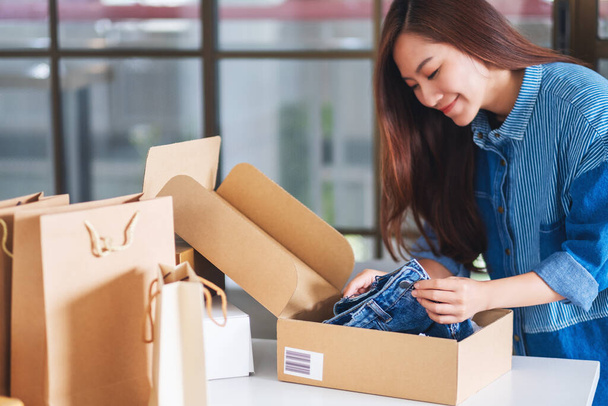 A beautiful young asian woman receiving and opening a postal parcel box of clothing at home for delivery and online shopping concept - Photo, Image