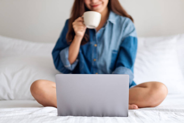 Κοντινό πλάνο εικόνα μιας όμορφης γυναίκας που χρησιμοποιεί και εργάζεται σε φορητό υπολογιστή, πίνοντας καφέ, ενώ κάθεται σε ένα λευκό άνετο κρεβάτι στο σπίτι - Φωτογραφία, εικόνα