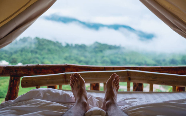 Zbliżenie obrazu stóp mężczyzny leżącego na białym, przytulnym łóżku w namiocie z pięknym widokiem na przyrodę w mglisty dzień na zewnątrz - Zdjęcie, obraz