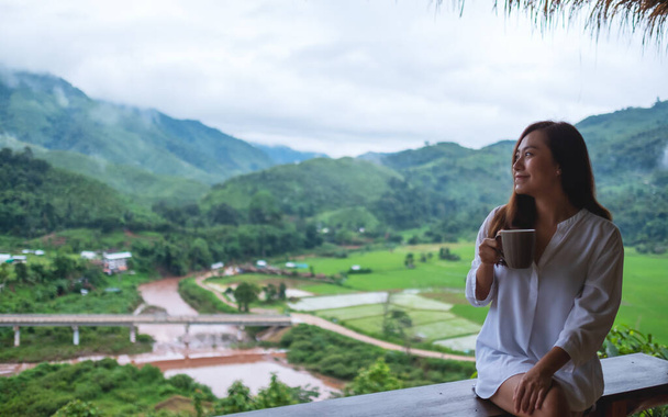 美しいアジアの女性が熱いコーヒーを飲み、バルコニーに座って山と緑の自然を見ている姿  - 写真・画像