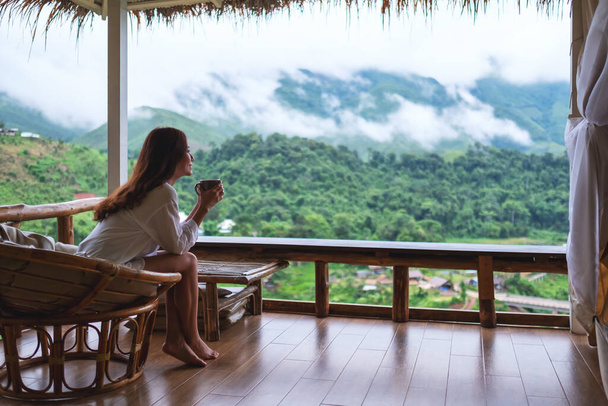 Portraitbild einer schönen asiatischen Frau, die heißen Kaffee hält und trinkt, auf einem Balkon sitzt und die Berge und die grüne Natur betrachtet  - Foto, Bild