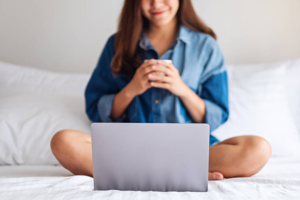 zbliżenie obraz z a piękny Azji kobieta za pomocą laptop komputer dla wideo rozmowy podczas siedzenie i picie kawa na a łóżko w domu - Zdjęcie, obraz