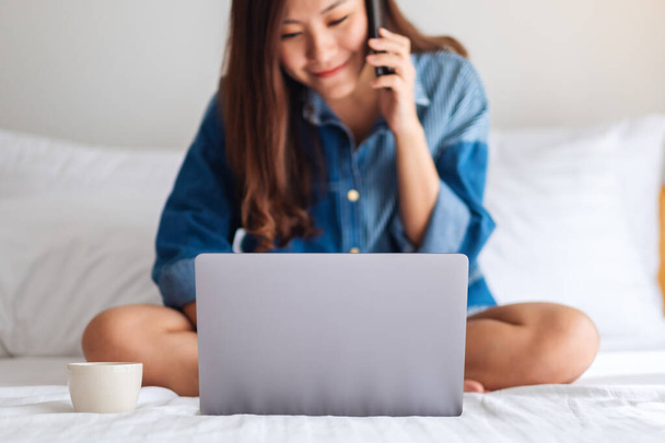Immagine ravvicinata di una bella giovane donna asiatica che usa e parla su cellulare e laptop mentre è seduta su un letto bianco - Foto, immagini