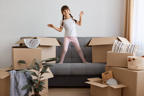 Indoor shot of happy smiling little girl wearing casual style attire standing on sofa and dancing, being happy to move into a new apartment, expressing positive emotions. - Foto, imagen