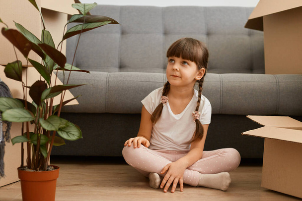 Portrait of charming dark haired little girl with braids wearing white t shirt sitting on floor near sofa and cardboard boxes, kid looking away with interest, admiring new house while relocating. - Фото, изображение