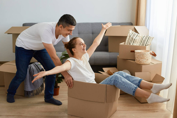 Full length portrait of positive couple is having fun with cardboard boxes in new house at moving day, husband riding his wife in carton box, expressing happiness. - Φωτογραφία, εικόνα