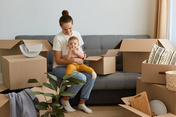 Image of Caucasian woman with bun hairstyle wearing white t shirt sitting on sofa with her infant baby, moving to a new flat, holding toddler daughter, needs to unpack personal belongings. - Photo, Image
