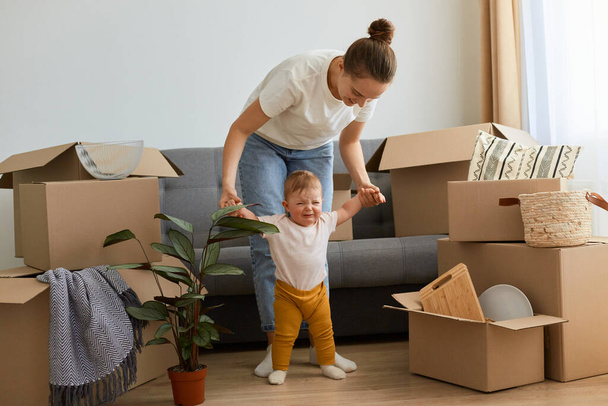 Portrait of woman wearing white t shirt standing near sofa with her infant daughter and holding her hands, teaching her cute child to do first steps, having fun while relocating into a new apartment. - 写真・画像