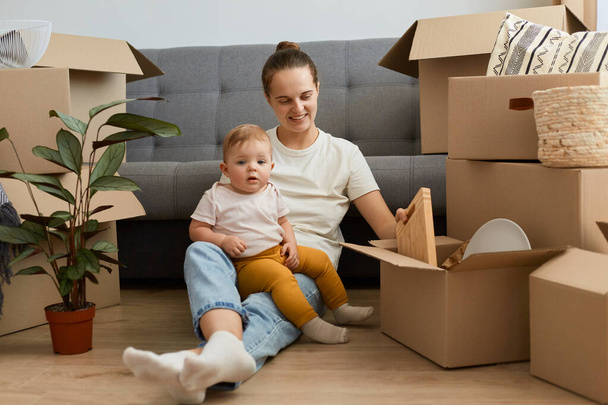 Indoor shot of smiling positive woman wearing white t shirt sitting on floor near sofa with her infant baby daughter, unpacking personal belongings, expressing happiness, moving day. - Foto, immagini