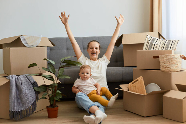 Full length portrait of excited satisfied happy woman wearing white t shirt sitting on floor near sofa with raised hands, rejoicing to relocate to a new house, holding her cute kid on legs. - Фото, зображення