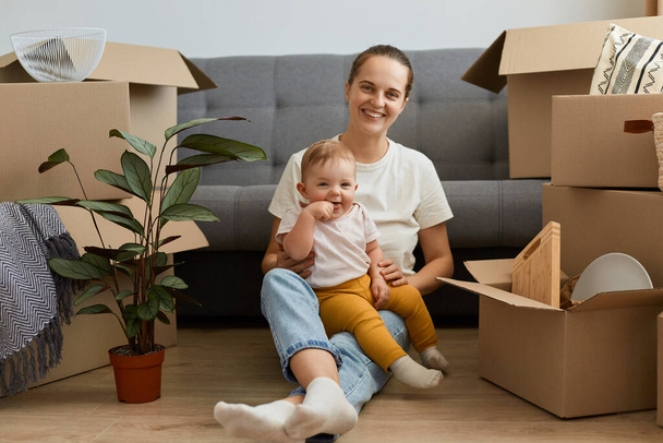 Portrait of satisfied woman wearing white t shirt sitting on floor near sofa, looking at camera with charming smile, holding her daughter, celebrating to move into a new house. - Фото, изображение