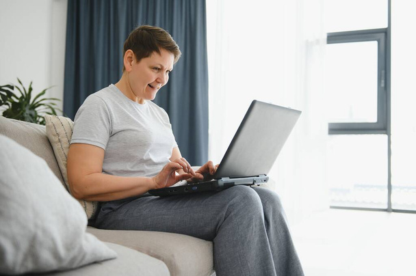 Technology, people and communication concept - Happy senior woman with tablet pc computer having video chat at home. Modern middle-aged 50s grandmother sit relax on couch in living room using laptop - 写真・画像
