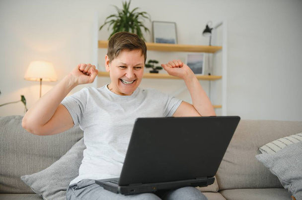 Happy excited grey haired mature woman celebrating online win, using laptop, looking at screen, sitting on couch at home, middle aged female feeling amazed, surprised by unbelievable good news - Фото, изображение