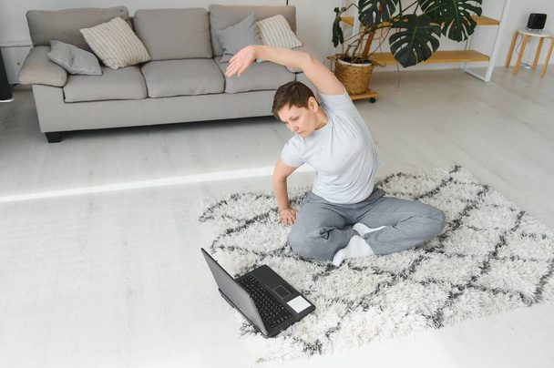 Fitness at home online. Middle aged woman in sportswear doing yoga on sports mat with laptop in living room interior with gadgets, profile, free space - Φωτογραφία, εικόνα