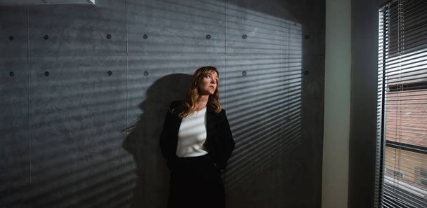A senior, brown-haired woman leans against gray, blind shadow background in a darkened room while she thoughtfully looks through the window. A formal clothed female professor stands next to the window. - Foto, Bild