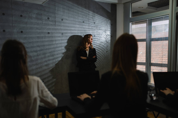 A middle-aged handsome female executive leans against the wall in a dark office and looks out the window as unrecognizable female employees sit at the table and look at her. - Photo, Image