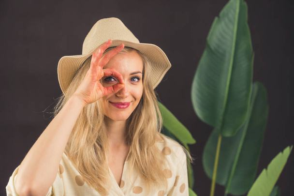 Śmieszny biały blondynka młoda dziewczyna stojąca przed duży-liść roślina w kapelusz robiący ok gest z ręka uśmiechając się, oko patrząc przez palce z szczęśliwy twarz. Koncepcja podróżnika. Safari. - Zdjęcie, obraz
