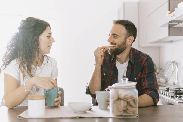Nişanlı çift yeni evlerinde kahvaltı yapıyor. Mutfakta içerken gülümseyen ve yemek yiyen genç çift. Arka planda sıcak bir filtre. - Fotoğraf, Görsel