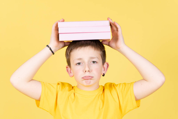 Netter gelangweilter Junge mit einem Stapel Bücher auf dem Kopf, isoliert auf gelbem Hintergrund. Stress und Angst bei Kindern. - Foto, Bild