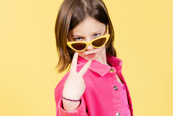 Menina arrogante usando óculos de sol posando para câmera isolada no fundo amarelo - Foto, Imagem