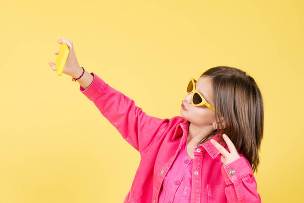 Arrogant and egocentric little girl wearing sunglasses posing and taking a selfie isolated on yellow background. - 写真・画像