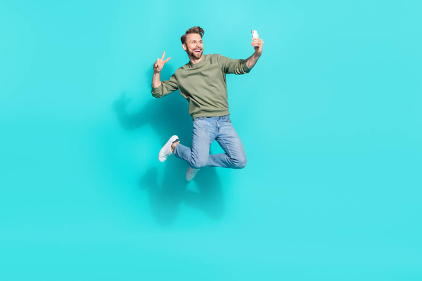 Full length profile photo of crazy blogger man jump hold telephone make selfie wear pullover isolated blue color background - Фото, зображення