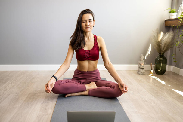 Fit woman doing yoga exercise while watching tutorial on laptop at home. Attractive chinese woman exercising on a mat and watching instructional videos on laptop. - Foto, Imagen