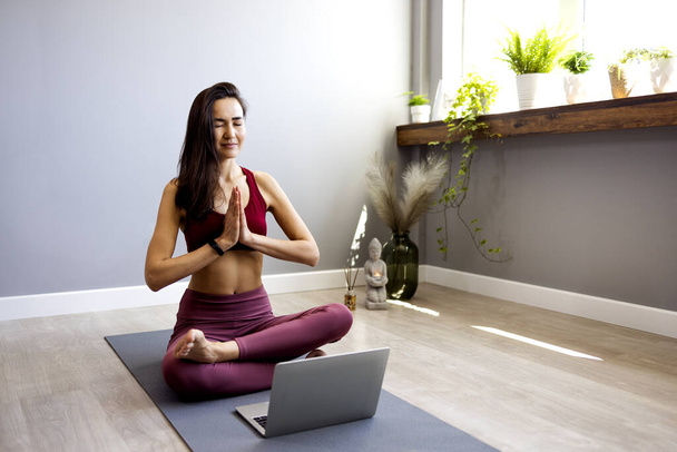 An Asian young sports woman is sitting on the floor in the living room, practicing yoga in an online computer class. A calm woman meditates at home watching classes on a webcam on a laptop. The concept of relaxation and yoga - Foto, afbeelding