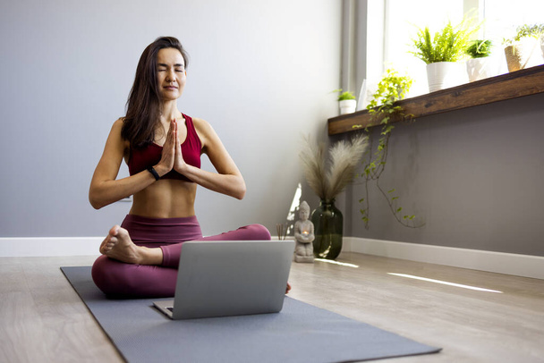 Uma jovem mulher esportiva asiática está sentada no chão na sala de estar, praticando ioga em uma aula de informática online. Uma mulher calma medita em casa assistindo aulas em uma webcam em um laptop. O conceito de relaxamento e ioga - Foto, Imagem