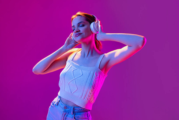 Portrait of young beautiful happy girl in headphones listening to music isolated on purple background in neon light, filter. Concept of emotions, music, facial expressions - Photo, image