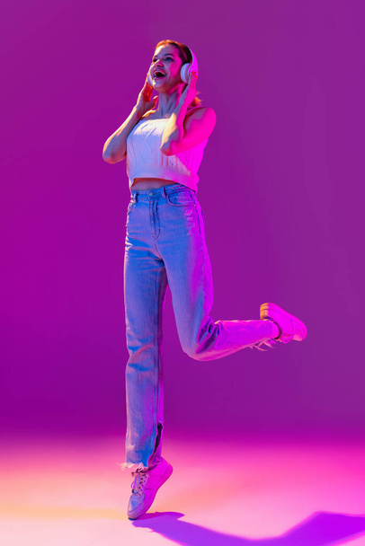 Young beautiful excited girl in headphones listening to music isolated on purple background in neon light, filter. Concept of emotions, music, facial expressions - Foto, Bild