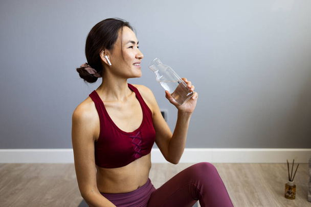 An Asian woman holds a bottle of water in her hand like a dumbbell as she finishes her workout at home. Women's sports lifting equipment for weight training. Wellness people, the concept of well-being - Foto, immagini