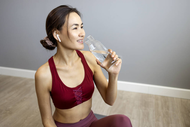 Asian beautiful woman in sportswear drink water after exercise at home. Thirsty active sport woman takes a sips of clean mineral natural in cup after workout for health care in kitchen in house. - Фото, изображение