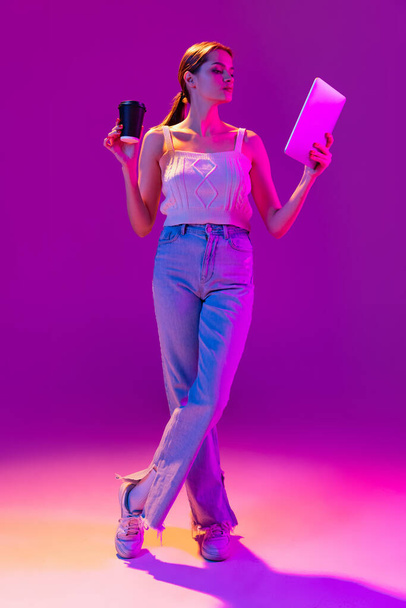 Young beautiful happy girl wearing casual style clothes using tablet isolated on purple background in neon filter. Concept of emotions, digital technologies, fashion - Fotografie, Obrázek