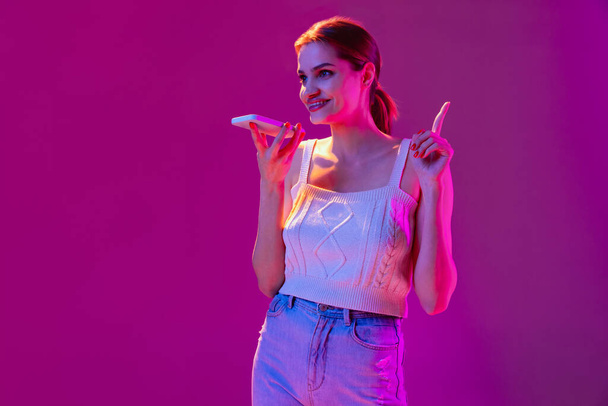 Young beautiful happy girl recording voice message on phone isolated on purple background in neon filter. Concept of emotions, digital technologies, fashion - Photo, Image