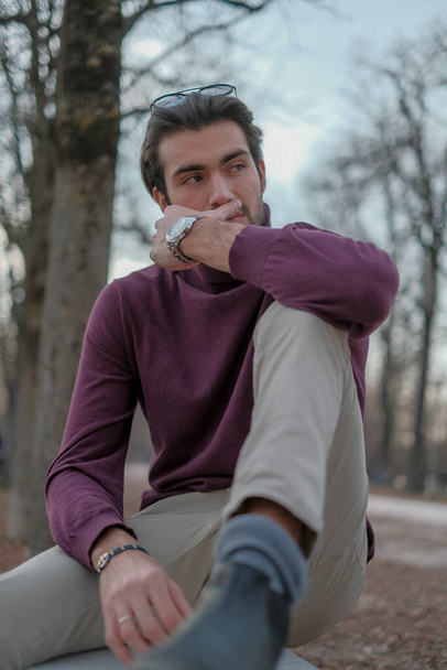 schöner italienischer dunkelhaariger Junge, der im Herbst mit dunkelrotem Pullover auf einer Bank sitzt. Hochwertiges Foto - Foto, Bild