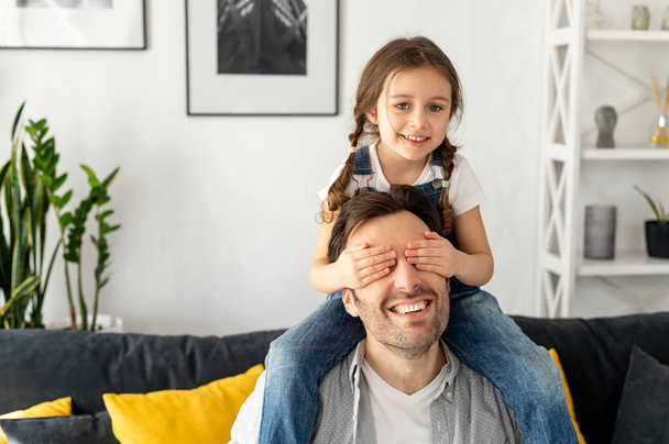 Cute girl closed eyes to her father. Loving father carrying his little toddler daughter on the shoulders sitting on the sofa in cozy living room, single parent playing with kid - Foto, Imagem