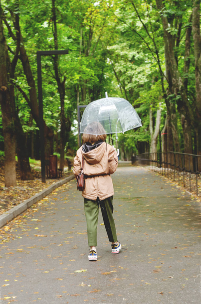 Young happy woman walking in the rain in green park with umbrella, smiling, fun. person sitting on the bench. Cold weather. Spring. Fall. - Zdjęcie, obraz