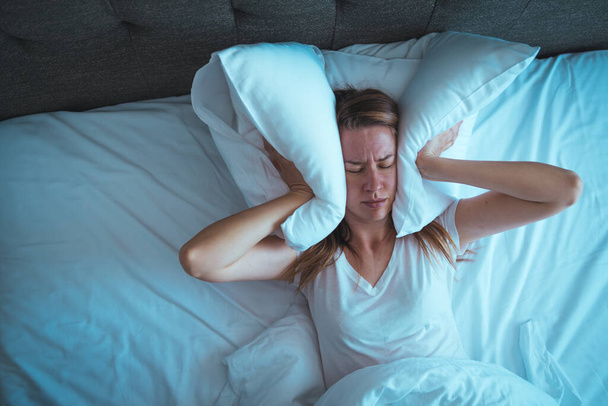 Young beautiful Caucasian woman on bed having headache / insomnia / migraine / stress. Young woman covering her face with hands. Desperate girl suffering insomnia trying to sleep - Photo, image