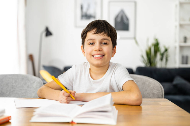 Lachende schooljongen die op afstand van huis studeert, knappe jongen die huiswerk maakt, taken schrijft in het notitieboekje, online lessen kijkt. E-studeren en thuisonderwijs concept - Foto, afbeelding