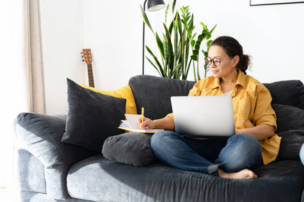 Focused woman freelancer researching business tasks, taking a notes with a laptop at home. Female is writing in a notebook, watching online classes, webinar - Foto, immagini