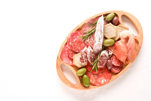 Wooden charcuterie board with different types of sausages - salami, bresaola, proscuitto served with olives and crackers. Traditional italian antipasti on white concrete table background with copy space. Selective focus - 写真・画像
