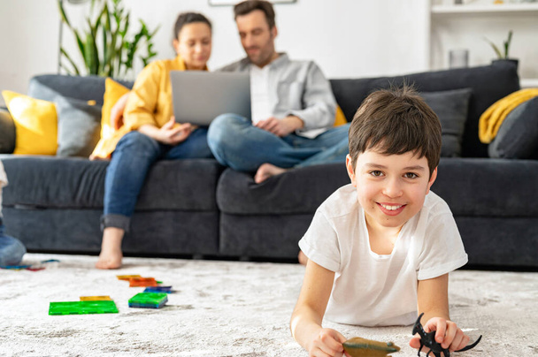 Two parents sitting on the sofa in cozy living room and looking at the laptop, while their son playing on the floor - Foto, imagen