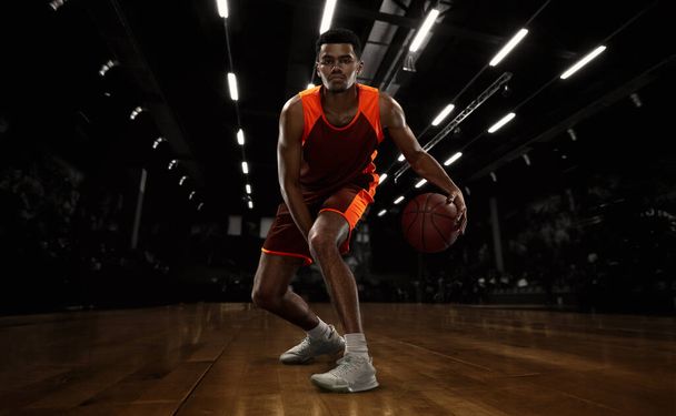 African-american young basketball player in action and motion in flashlights over dark gym background. Concept of sport, energy and dynamic, healthy lifestyle. Arenas drawned - Foto, afbeelding