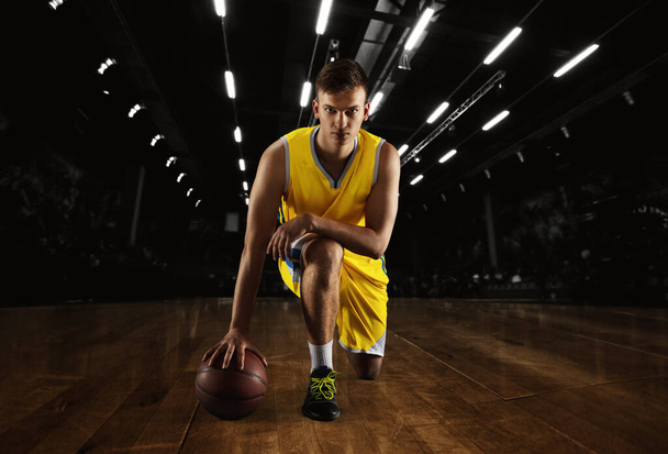 Young caucasian male basketball player posing with ball isolated over dark gym background with flashlights. Concept of motion, power, speed, healthy lifestyle, professional sport. - Photo, Image