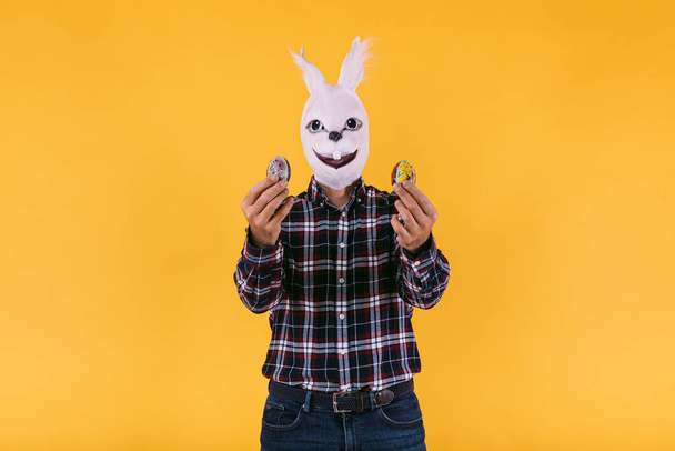 Persona disfrazada con máscara de conejo con camisa a cuadros y jeans, sosteniendo huevos de Pascua, sobre fondo amarillo. Carnaval, fiesta, pascua y concepto de celebración. - Foto, Imagen